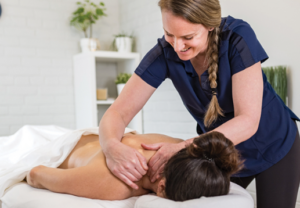 An RMT with long blond braided hair, wearing a blue scrub top, is giving a massage treatment to a person's shoulders and neck. The person is lying prone on a massage table.