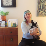Kirby a white woman in her late 30s with medium-length green hair, is sitting in her massage room, smiling and holding her one employee, Wilma the blind boston terrier. 