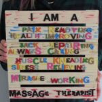 Kirby holds a small wooden pallet sign with colorful letters. A gifted, created by her by her mother. The sign reads I am a pain-removing, health-improving, back-repairing, always caring, muscle-kneading, stress-relieving, miracle-working, Massage Therapist.