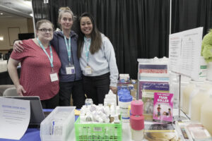 Three people standing together at a trade show booth. There are massage therapy supplies on the table in front of them.