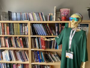 Skeleton Seemore Bones wearing a green t-shirt, tie-dye bandana and reflective sunglasses. Seemore is standing beside a bookcase in the MTAM Resource library.