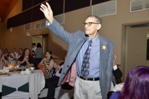 An elderly gentleman (Peter Stachnyk) is standing and waving in thanks as his seated colleagues applaud around him.