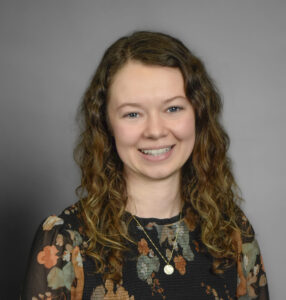 Headshot of Samantha Friesen. A smiling woman with long, light brown balayage, curly hair. She is wearing a black long sleeved floral patterned blouse and silver necklace.