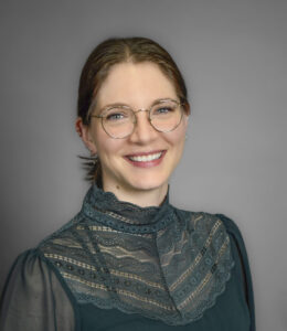 Headshot of Natashia Durand. A smiling woman with brown hair pulled back. She has thin gold rimmed circular glasses and is wearing a dark green lace blouse.