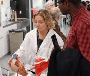A woman with blond hair wearing a white lab coat and holding an anatomical model of a human heart.